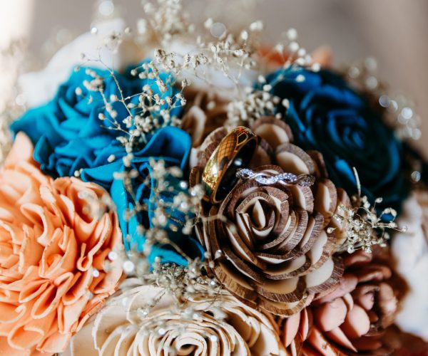 A close-up of a colorful bouquet with rings resting on paper flowers, featuring vibrant blue and peach hues, accented by delicate white blossoms.