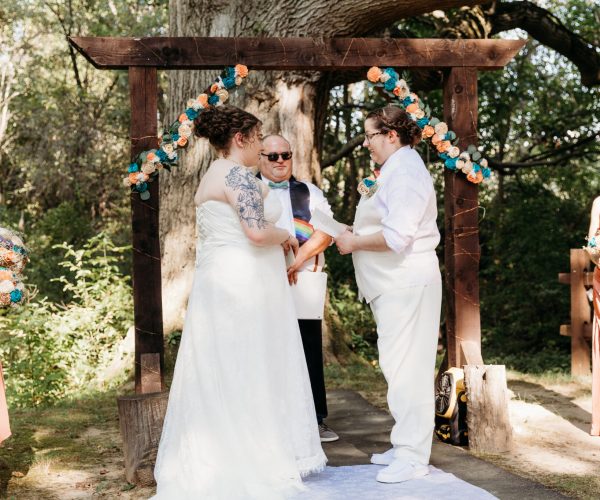 Two people exchange vows under a decorated wooden arch in a forest setting, with attendants holding bouquets and a person officiating.