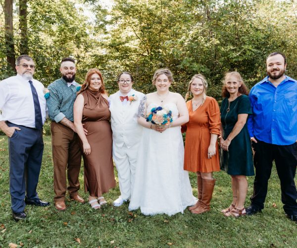 A group of eight people dressed in formal attire stand outdoors on grass, with trees in the background, appearing happy and relaxed.
