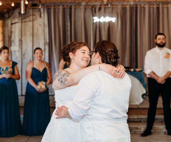 Two people dance at a wedding reception with onlookers dressed in blue. String lights hang overhead in a rustic venue setting.