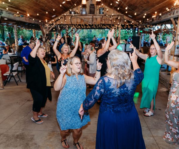 A group of people happily dancing at a rustic venue adorned with string lights and wooden beams, creating a lively celebratory atmosphere.