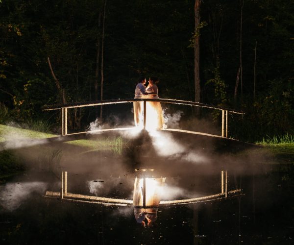 Two people stand on a wooden bridge over a misty pond, surrounded by trees at night, creating a romantic atmosphere.