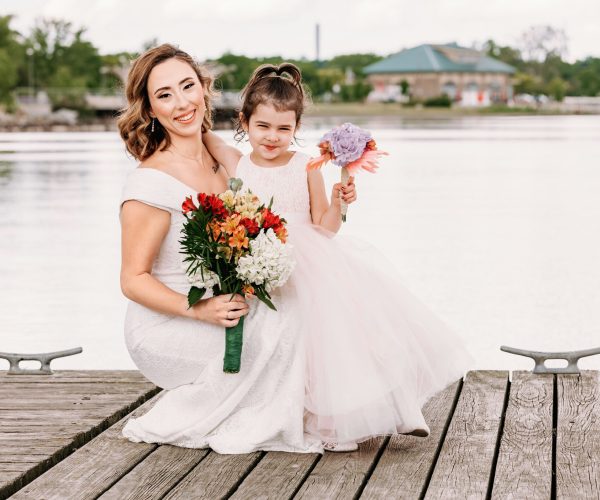 A person in a white dress and a young girl in a tulle skirt hold bouquets on a wooden dock by the water, smiling joyfully.