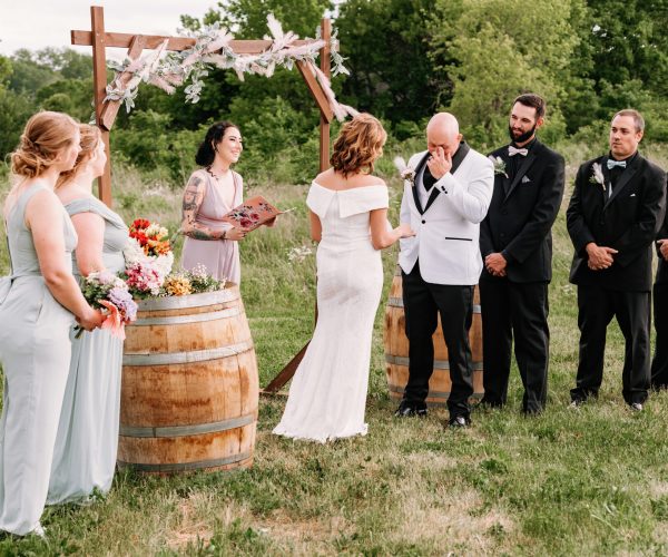 A wedding ceremony outdoors with a person in a white dress, person wiping tears, and attendants wearing gray and black suits beside a floral arch.