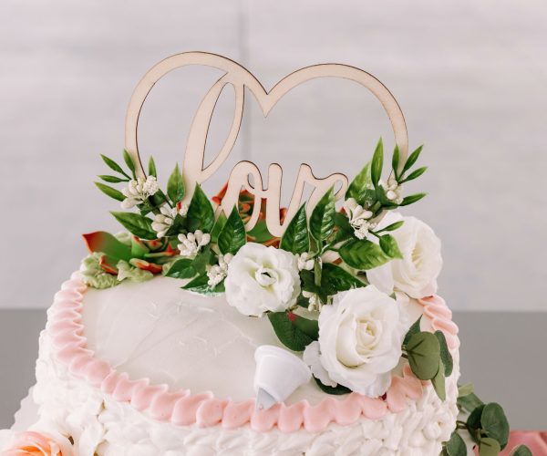 The image shows a cake with pink ruffles, white frosting, and a topper with two hearts and the word "love," adorned with white flowers and greenery.
