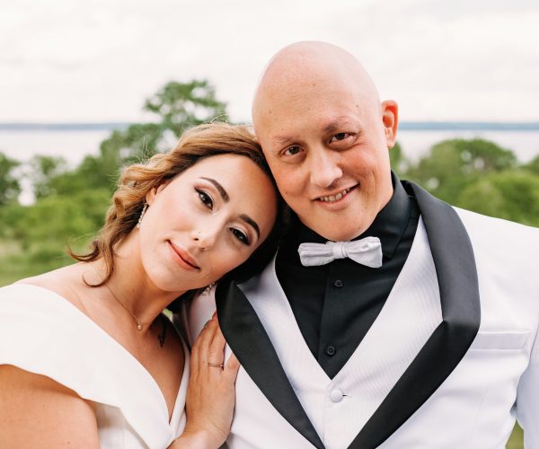Two people in formal attire smile outdoors, possibly a wedding scene, with trees and a lake in the background. They appear happy and content.