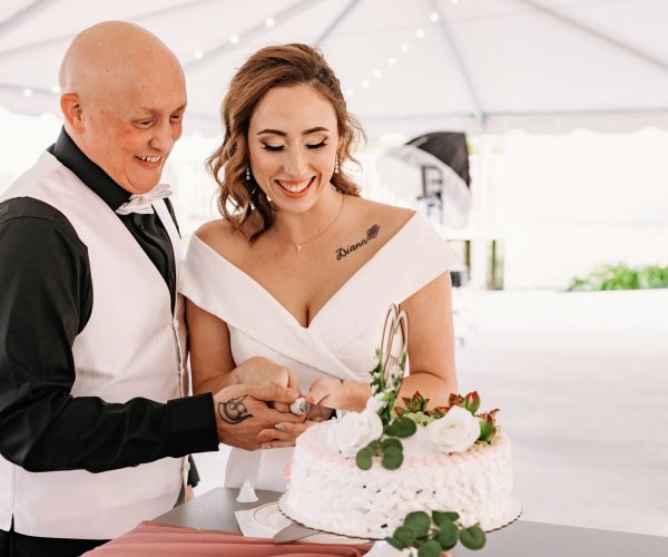 Two people dressed formally, presumably a couple, are happily cutting a white frosted cake adorned with flowers at an event decorated with string lights.