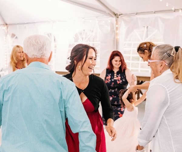 A group of people are happily interacting at an event under a white tent with string lights. A person in a red dress is smiling joyfully.