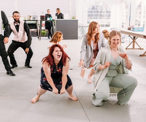 A group of people is smiling and dancing at an outdoor event near a white tent, with two individuals crouching at the forefront having fun.