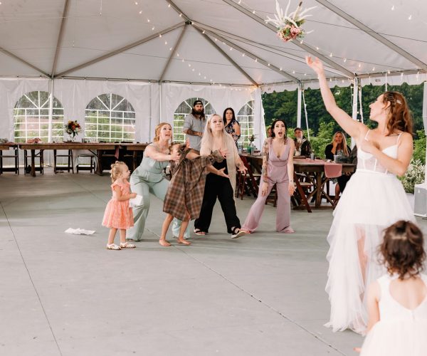 Guests excitedly reach out to catch a bouquet tossed by a bride under a tent at a wedding reception; a moment of joyful anticipation captured.