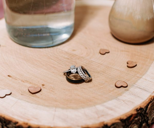 Three rings, possibly wedding bands and an engagement ring, are stacked on a wooden surface with small heart-shaped decorations and vases nearby.