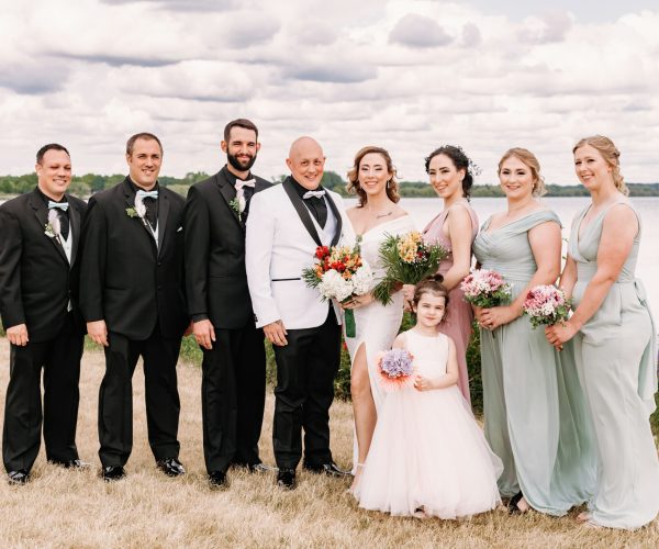 A wedding group photo by a lakeside with seven adults dressed formally and a child holding flowers, under a cloudy sky. They are smiling.