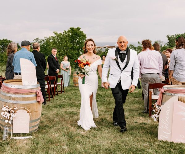A person in white attire walks down an aisle with a smiling person in a wedding dress, both looking joyful amid guests standing at an outdoor ceremony.