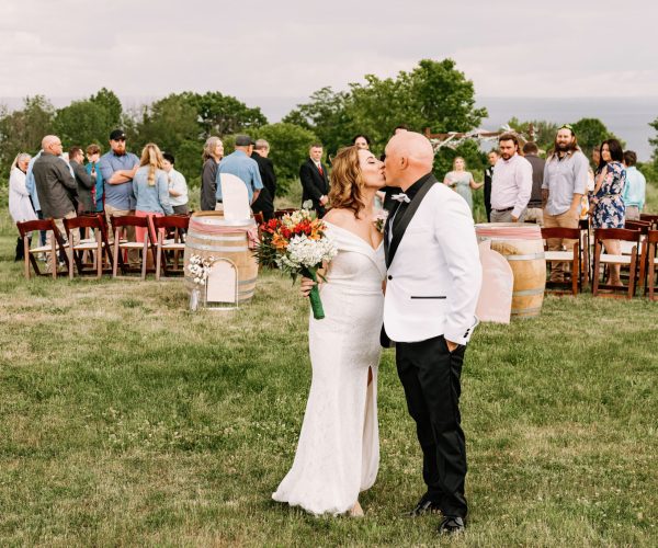 A person and a person in wedding attire share a moment among guests at an outdoor ceremony with open field and cloudy sky.