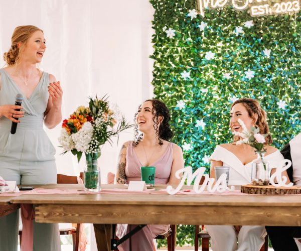 A person is giving a speech at a wedding reception, holding a microphone. The newlyweds and a guest are smiling at the table, with a "Mrs & Mrs" sign.
