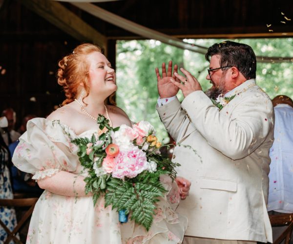 Two people celebrate at a wedding, surrounded by guests. One holds a floral bouquet, smiling joyfully amid a festive atmosphere.